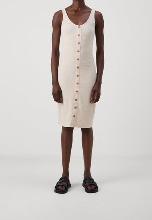 Beige ærmeløs ribbet kjole med knapper foran, båret med sorte slidssandaler på en mørkhudet person, der står mod en ensfarvet baggrund.