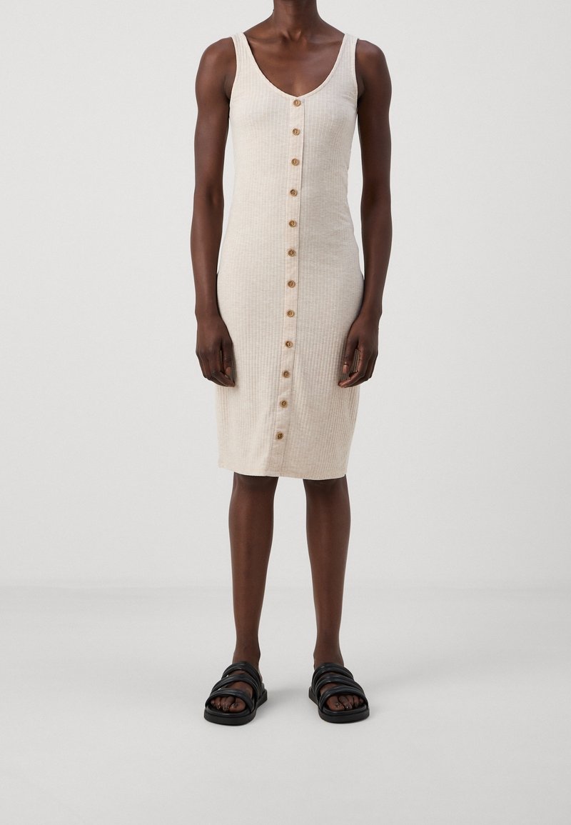 Robe beige sans manches côtelée avec des boutons devant, portée avec des sandales noires à enfiler sur une personne à la peau foncée, debout devant un fond uni.