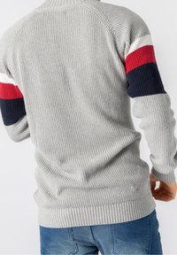 Šedý pletený svetr s ribovanou texturou, který má červené a modré pruhované akcenty na rukávech. Úzký střih a kulatý výstřih.