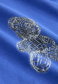 Zils kokvilnas audums ar teksturētu melnu lāču grafiku ar rakstu dizainu. Ievērojamas detaļas ietver bumbu ar burtiem 'B'.