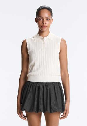 Mladá žena so zatvorenými vlasmi, oblečená v bezrukávovom bielom pletenom polotriku a čierno-bielej károvanej sukni.