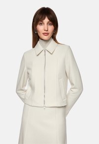 White wool-blend jacket with a pointed collar, front zipper closure, side pockets, and structured fit, paired with a matching skirt.