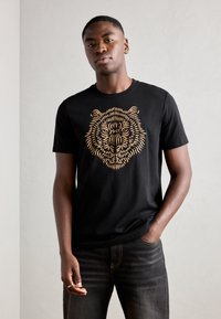 Black cotton t-shirt featuring a detailed golden tiger graphic on the front. Round neckline and short sleeves; worn with dark jeans.