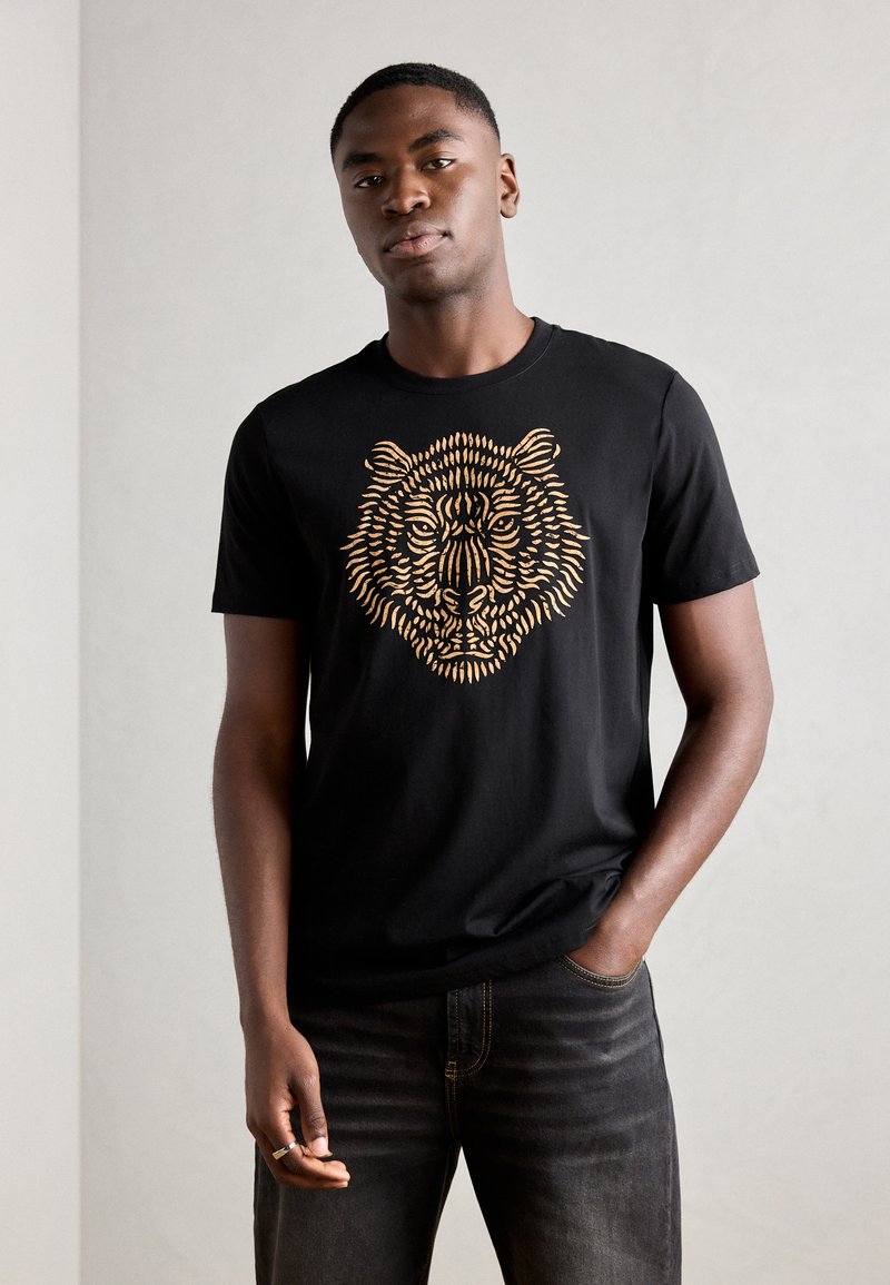 Camiseta de algodón negra con un gráfico detallado de un tigre dorado en la parte frontal. Cuello redondo y mangas cortas; combinada con vaqueros oscuros.
