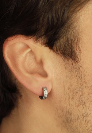 Texturierter silberner Hänger auf einem Ohr, der ein geometrisches Muster und ein kompaktes Design aufweist. Minimalistischer Stil mit glattem Finish.