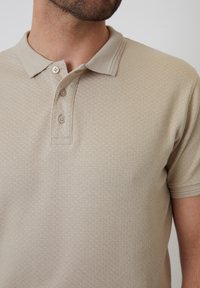 Beige polo tričko vyrobené z texturované tkaniny, které má límeček a tři knoflíky na zapínání. Krátké rukávy s jemným vzorem.