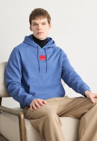 Hettegensere i lyseblå bomull med en frontlomme og et rødt logopatch. Modellen har på seg svart høyhalset genser og beige bukser, sittende.