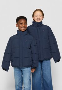 Marina puffjackor med höga kragar, med stora kviltade sektioner och logotypdetaljer, som bärs över denimjeans av två barn.