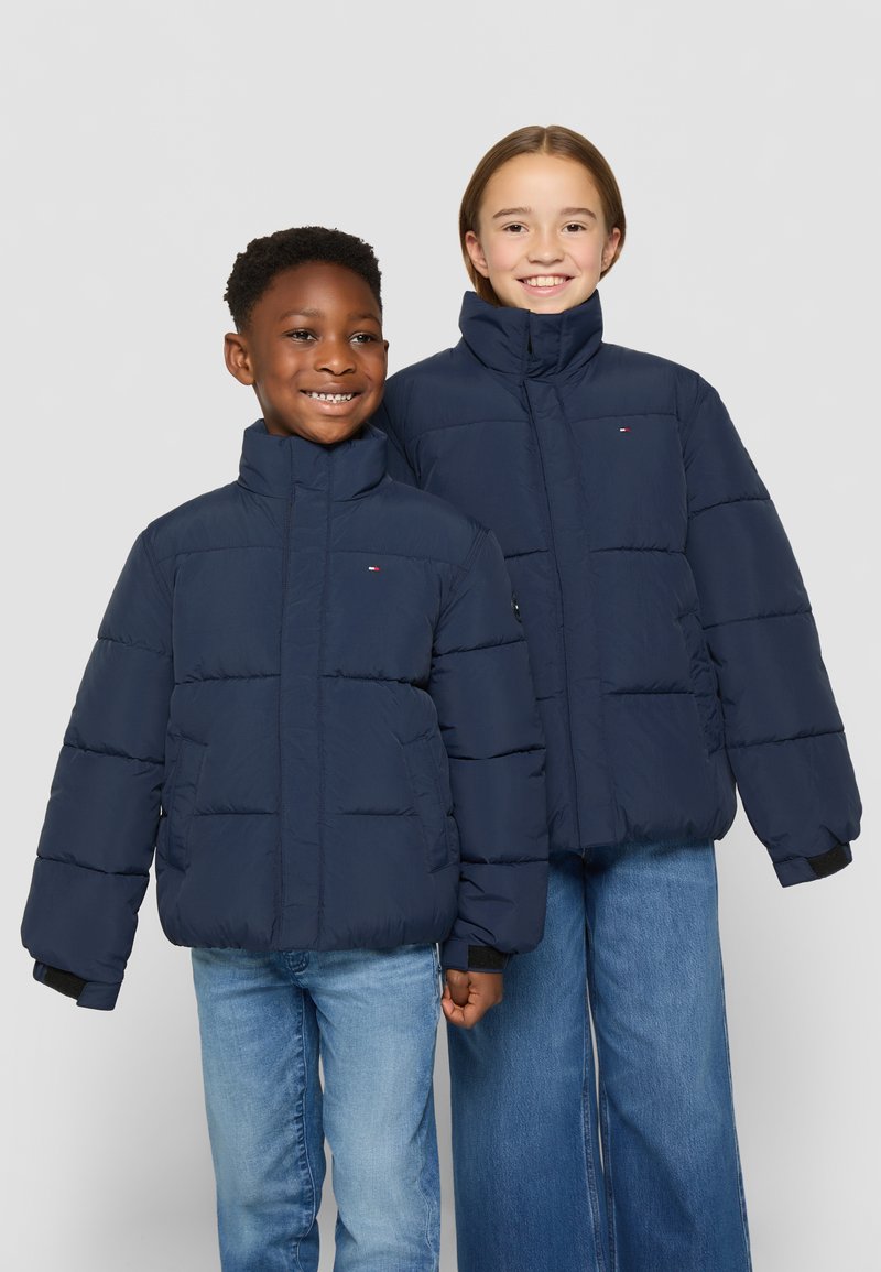 Marina puffjackor med höga kragar, med stora kviltade sektioner och logotypdetaljer, som bärs över denimjeans av två barn.