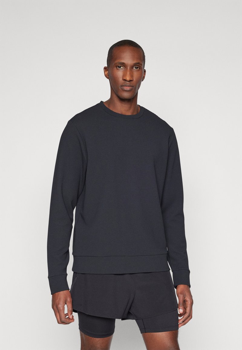 Mørkegrå sweatshirt med teksturert overflate, rund hals og lange ermer, brukt over svarte korte bukser. Enkel, minimalistisk design.