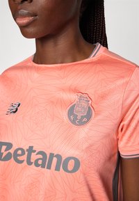 Heled roosa spordijakk geomeetriliste mustritega, hallide aktsentide ja ümmarguse logoga. Omab ümarat kaelust ja lühikesi varrukaid.