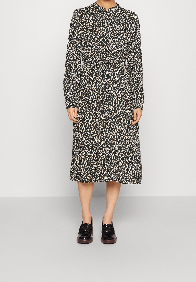 Woman wearing a black midi dress with beige and white abstract spots, long sleeves, button-up front, and tied waist, paired with black loafers.