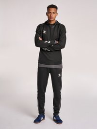 Svart träningshuva med texturerad axeldesign, i kombination med matchande svarta byxor med sidostråk. Blå träningssko fulländar outfiten.