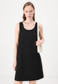 Svart ärmlös klänning med rundad halsringning, tillverkad av mjukt tyg; modellen håller en liten beige handväska. Enkel, A-linjeformad silhuett, minimal design.