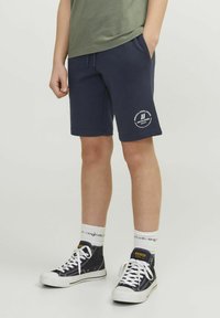 Pantalones cortos de algodón azul marino con bolsillos laterales, que presentan un logo impreso en blanco. Combinados con zapatillas altas negras y calcetas blancas acanaladas.