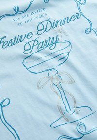 Einladung zur festlichen Dinnerparty mit einem blauen Champagnerglas und silbernem Band auf einem hellblauen Stoffhintergrund.