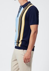 Man in een marineblauwe polo met korte mouwen, met blauwe en gele verticale strepen, gecombineerd met beige broek tegen een witte achtergrond.