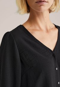 Camicia nera a bottoni con tessuto testo, design a V e maniche leggermente gonfie. I bottoni sono rotondi e posizionati lungo la parte anteriore.