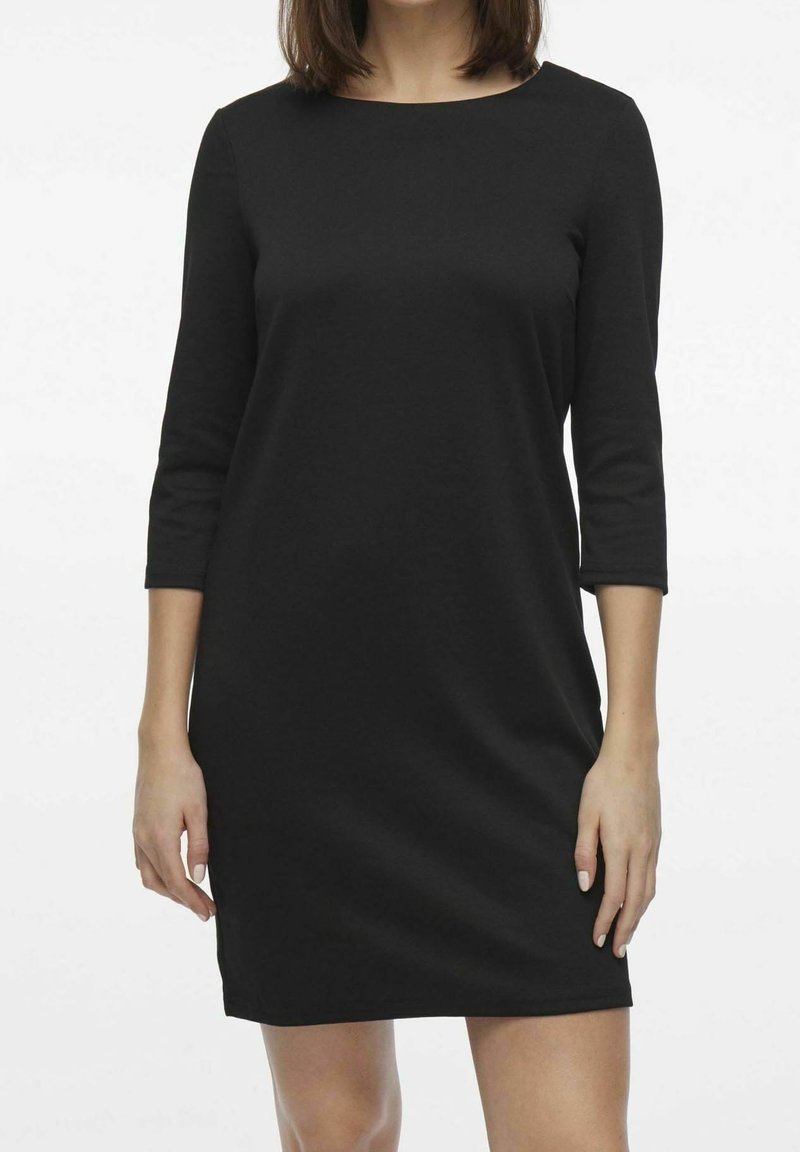 Femme portant une robe noire unie longueur genoux avec des manches trois-quarts, debout devant un fond blanc.