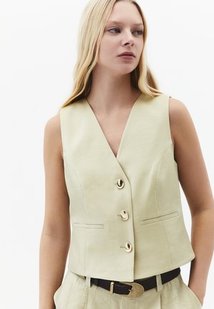 Kvinde med langt blondt hår iført en ærmeløs beige vest med guldknapper og matchende bukser med sort bælte mod en hvid baggrund.