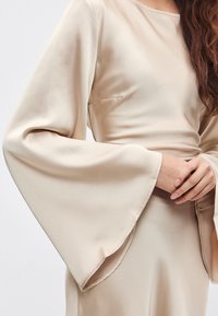 Smėlio spalvos satino palaidinė su ilgomis, plačiomis rankovėmis ir valtine iškirpte, pasižyminti elegantišku paviršiumi ir minkšta, lygiu tekstūrų.