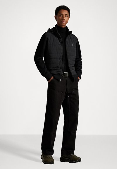 Μαύρο επενδυμένο γιλέκο με κουκούλα πάνω από μαύρο φούτερ με ψηλό λαιμό, συνδυασμένο με μαύρες παντελόνες φαρδιάς γραμμής και σκούρα πράσινα αθλητικά παπούτσια, με λογότυπο στο γιλέκο.