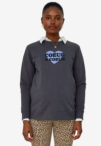 Gray sweatshirt with round neckline, featuring a heart graphic and "COEUR À COEUR" text. Worn over a white collared shirt and leopard print pants.