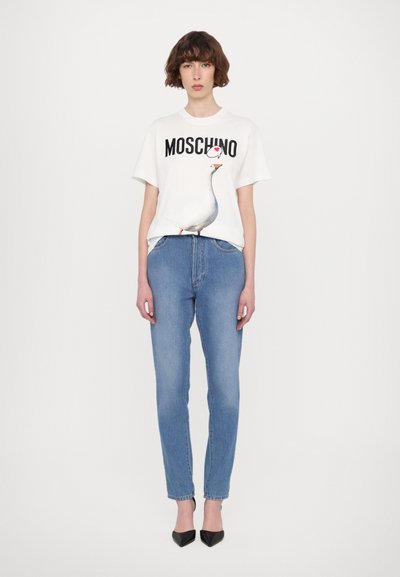 Balta marškinėliai su "Moschino" užrašu ir žąsies grafika, derinami su šviesiai mėlynomis džinsinėmis kelnėmis ir juodais smailiais kulnais.