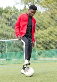 Chaqueta roja de Nike sobre una camiseta negra, pantalones negros con rayas blancas y zapatillas blancas; una persona equilibra un balón de fútbol sobre su pie en el césped.