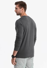 Langärmeliges graues Shirt mit Rundhalsausschnitt, schmaler Passform und glatter Textur. Verfügt über ein dezentes Markenlabel am Saum. Kombiniert mit hellen Cargo-Hosen.