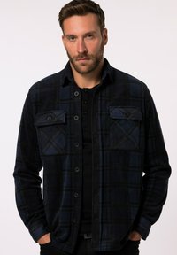 Marineblauw en zwart geruite flanellen shirt met knoopzakken op de borst, volledige knoopsluiting aan de voorkant en een zachte textuur. Gestyled over een zwart T-shirt.