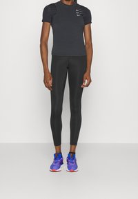 Camiseta deportiva negra con detalles texturizados en los hombros, combinada con leggings negros y zapatillas azul-púrpura con acentos rojos.