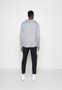 Man in een grijze t-shirt met lange mouwen, zwarte sportbroek met witte strepen, witte sokken en witte sneakers, staande met zijn gezicht naar een witte muur.