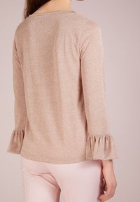 Haut rose pâle scintillant avec manches trois-quarts et poignets à volants. Le tissu a une texture lisse et un col rond simple.