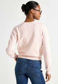 Maglione rosa chiaro con accenti di pompon bianchi testurizzati, polsini e orlo a coste, abbinato a jeans in denim blu a vita media.