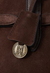 Braune Wildledertasche mit Details, die ein kreisförmiges goldenes Anhänger mit geprägt Design und Text zeigt. Sie enthält Lederakzente und kontrastierende Nähte.