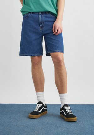 Άτομο που φοράει μπλε τζιν σορτσάκι, λευκές κάλτσες πλήρους μήκους και μαύρα αθλητικά παπούτσια με λευκά κορδόνια σε μπλε χαλί.