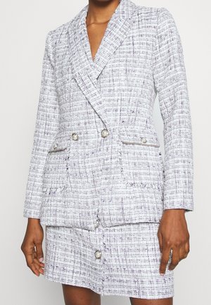 Frau trägt einen weiß-grau karierten Tweed-Blazer und passenden Rock mit perlenähnlichen Knöpfen und Fransen-Details.