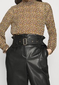 Langärmlige Bluse mit hohem Kragen in Braun mit einem floralem Muster aus gelben, orangefarbenen und roten Blumen; kombiniert mit hoch taillierten schwarzen Lederhosen mit einem breiten Gürtel.
