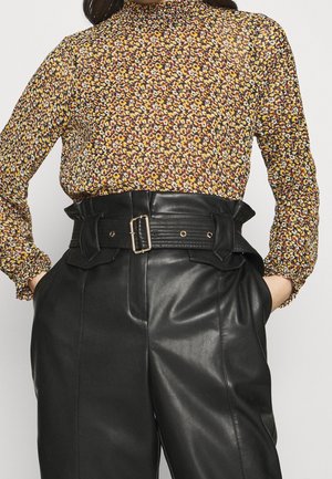 Langärmlige Bluse mit hohem Kragen in Braun mit einem floralem Muster aus gelben, orangefarbenen und roten Blumen; kombiniert mit hoch taillierten schwarzen Lederhosen mit einem breiten Gürtel.