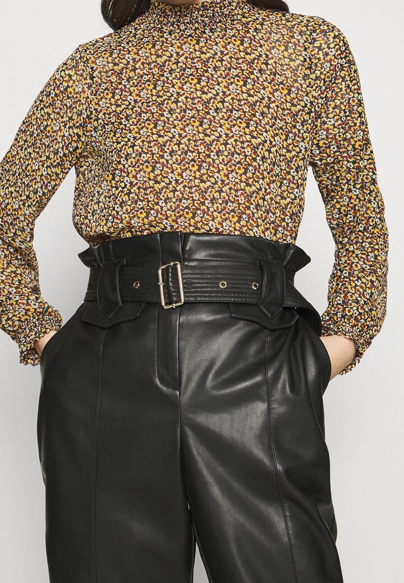 Langärmlige Bluse mit hohem Kragen in Braun mit einem floralem Muster aus gelben, orangefarbenen und roten Blumen; kombiniert mit hoch taillierten schwarzen Lederhosen mit einem breiten Gürtel.