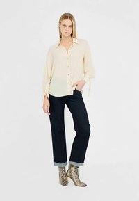 Kreemikas nööbitav pluus pikkade varrukatega ja lipsudetailidega, kantud tumedate teksade ja läikivate madu mustriga pahkluu saabastega.