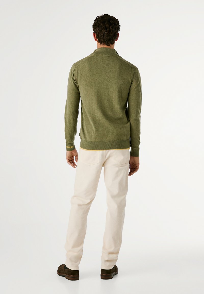 Mężczyzna stojący tyłem, ubrany w oliwkowy sweter, jasnobeżowe spodnie i ciemnobrązowe buty na białym tle.