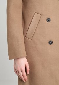 Brun yllekappa med slagkrage, dubbelknäppt design och två knappar. Vänster arm visar en stängd ficka och en slät textur.