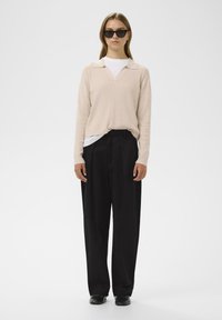 Beige, gerippter Strickpullover mit V-Ausschnitt und Kragen, über einem weißen T-Shirt getragen. Schwarze, weit geschnittene Hose und schwarze Schuhe komplettieren das Outfit.