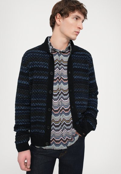 Adīta džemperis tumši zilā un melnā krāsā ar zigzaga rakstu. Iespējas ar pogām un apkakli. Valkāts virs rakstainas krekla un džinsiem.