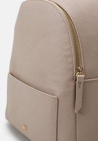 Mochila de cuero sintético beige con forma redondeada, con cremallera dorada y un bolsillo frontal. Superficie texturizada con detalles de patrón sutiles.