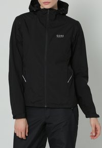 Persona che indossa una giacca con cappuccio Gore Running Wear nera e pantaloni neri, in piedi davanti a uno sfondo grigio chiaro uniforme.