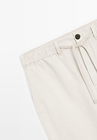 Beige cotton shorts with an elastic waistband, featuring a drawstring, a single black button, and side pockets. Smooth texture.