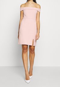 Femme portant une robe ajustée légère rose pâle à épaules nues avec une fente à l'avant, accompagnée de chaussures à talons pointus blanches, se tenant devant un fond uni.